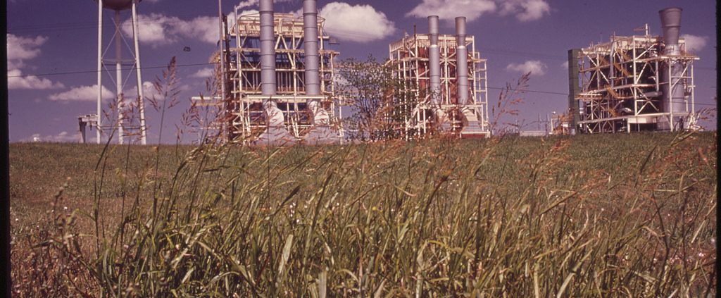 An image of pipes and smokestacks stretching up into a blue sky from a large field of grass.