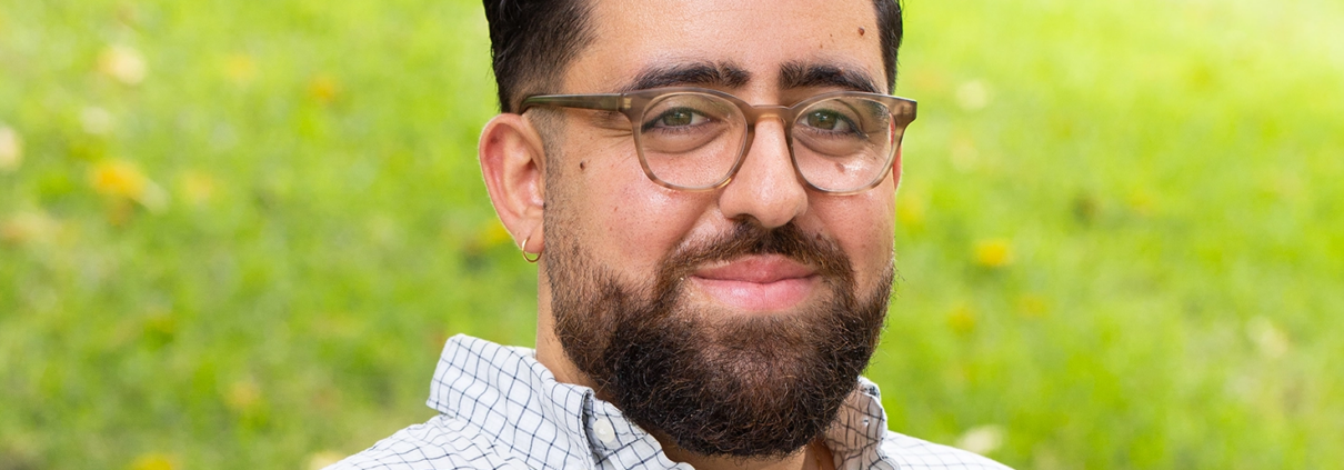 Portrait of Chris Jadallah outdoors on a grassy lawn, smiling at the camera. He has short dark curly hair, glasses, a beard, and is wearing a light checkered button-down shirt. The background is softly blurred greenery.