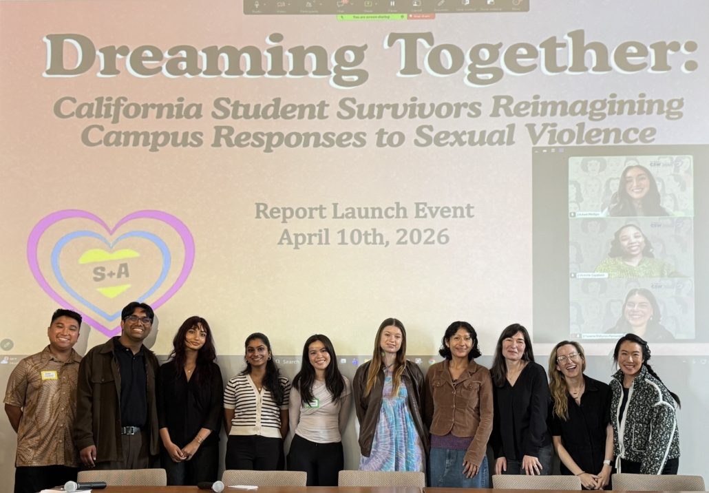 A group of ten people stand in a line on a stage, smiling for a photo. They are positioned in front of a long, dark wood table and a large projector screen. The screen displays the title "Dreaming Together: California Student Survivors Reimagining Campus Responses to Sexual Violence," along with the text "Report Launch Event April 10th, 2026." On the right side of the screen, a video call window shows three additional people participating remotely. The room has high ceilings, light-colored walls, and a wooden podium to the left of the group.