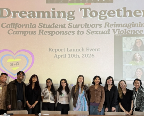 A group of ten people stand in a line on a stage, smiling for a photo. They are positioned in front of a long, dark wood table and a large projector screen. The screen displays the title "Dreaming Together: California Student Survivors Reimagining Campus Responses to Sexual Violence," along with the text "Report Launch Event April 10th, 2026." On the right side of the screen, a video call window shows three additional people participating remotely. The room has high ceilings, light-colored walls, and a wooden podium to the left of the group.