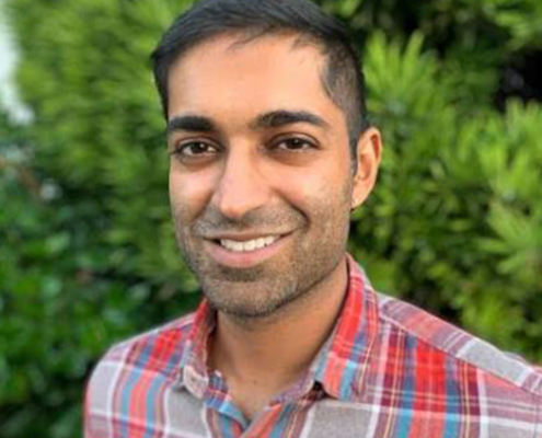 Portrait of Bharat Venkat. Man with short dark hair and checkered shirt standing in front of green bushes.