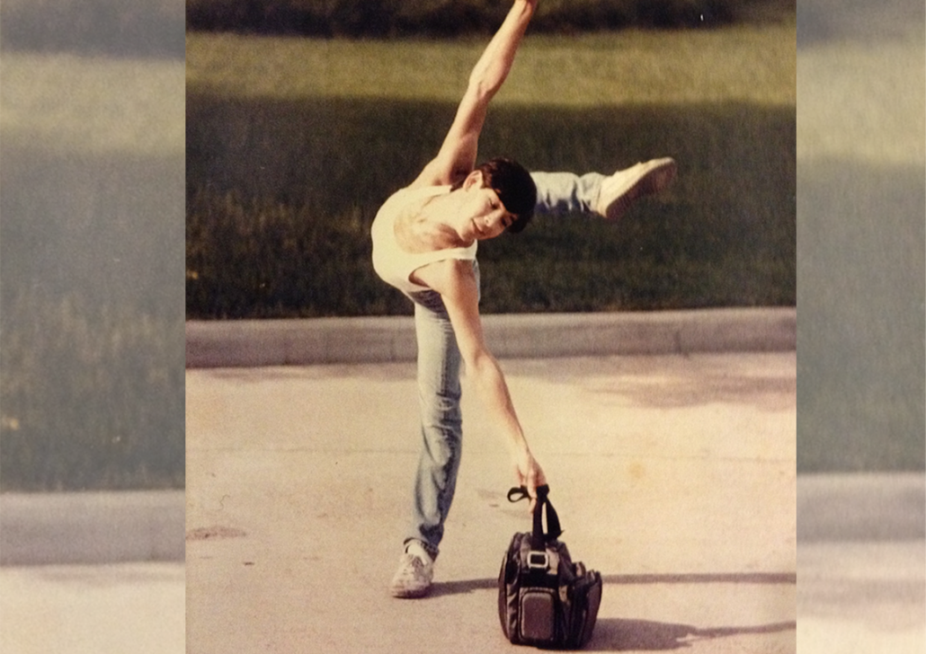 Carlos_Guzman A person balances on one leg with one arm and leg extended, reaching toward a duffel bag on the ground, outdoors on a paved surface.