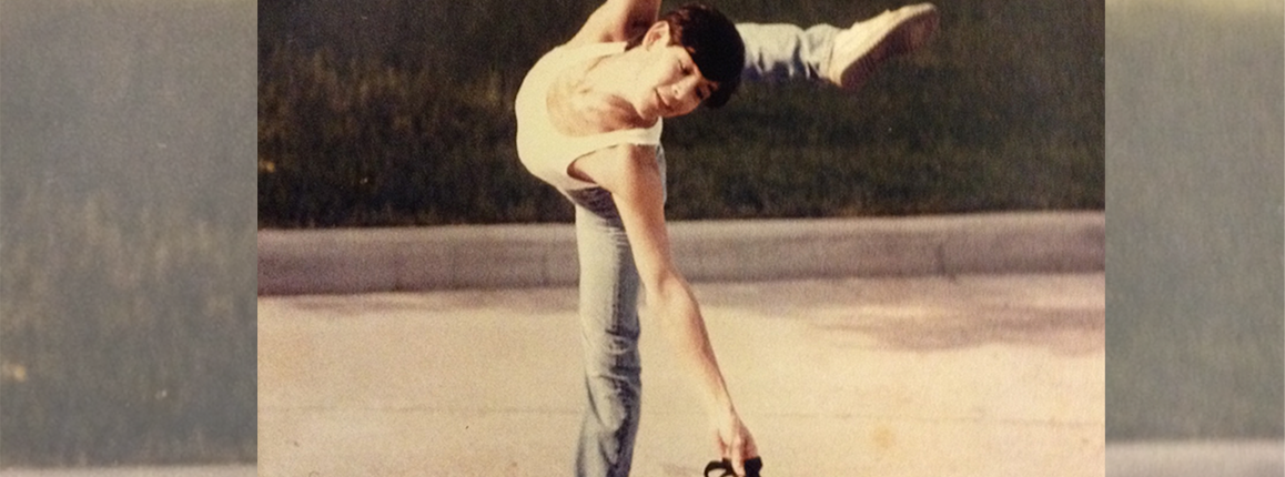 A person balances on one leg with one arm and leg extended, reaching toward a duffel bag on the ground, outdoors on a paved surface.