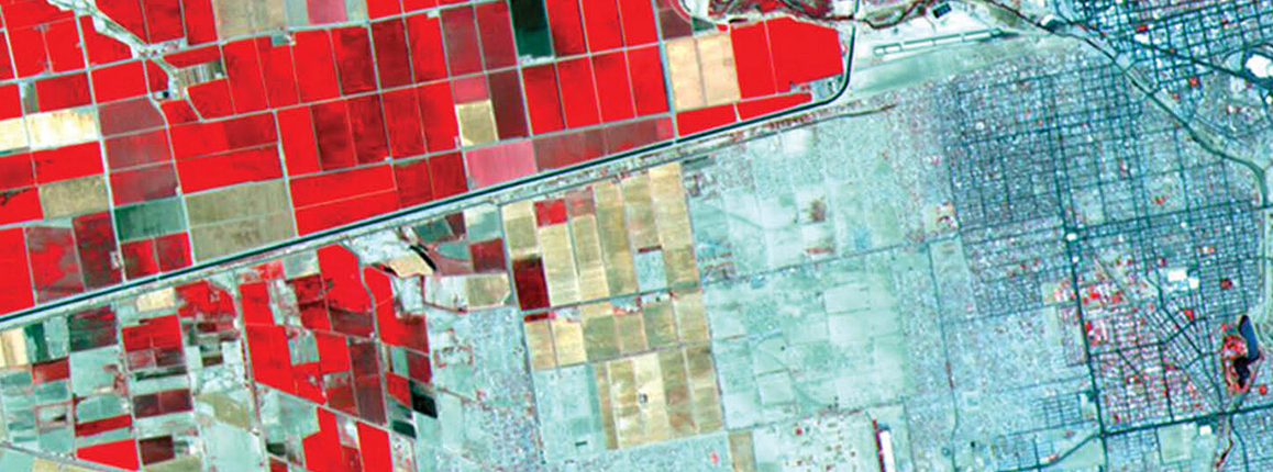 False-color satellite image showing a patchwork of rectangular agricultural fields in bright shades of red, dark red, tan, and green on the left, contrasted with a dense gray-blue urban grid on the right. A winding river runs across the upper portion of the image, while straight roads and canals divide the farmland into geometric blocks. The red coloration indicates healthy vegetation in infrared imagery.