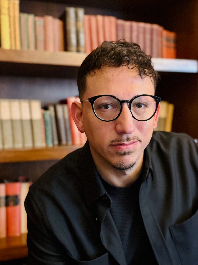 Joshua Guzman_headshot A person with short curly hair, glasses, and a goatee wears a dark shirt and sits in front of bookshelves filled with books.