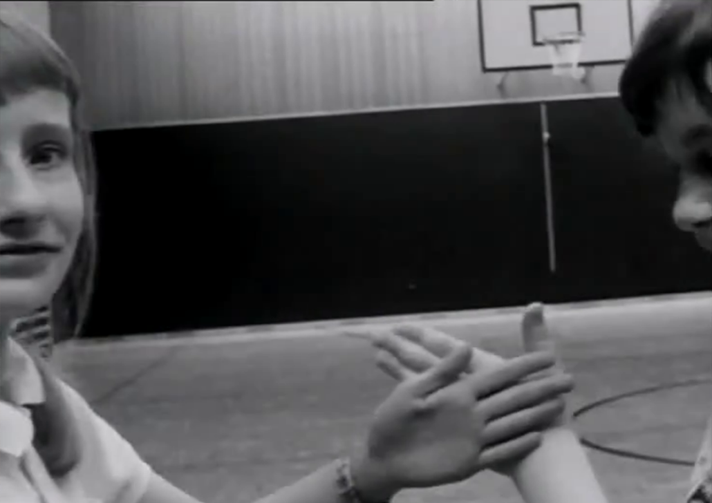 KalaniMitchell_Project Black-and-white photograph of two children facing each other with their hands raised as if playing a hand-clapping game in a gymnasium. A basketball hoop is visible in the background.