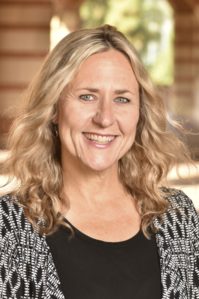 Lené Levy-Storms headshot Image of smiling blond person with curly shoulder length hair. The headshot is take in front of brick buildings.