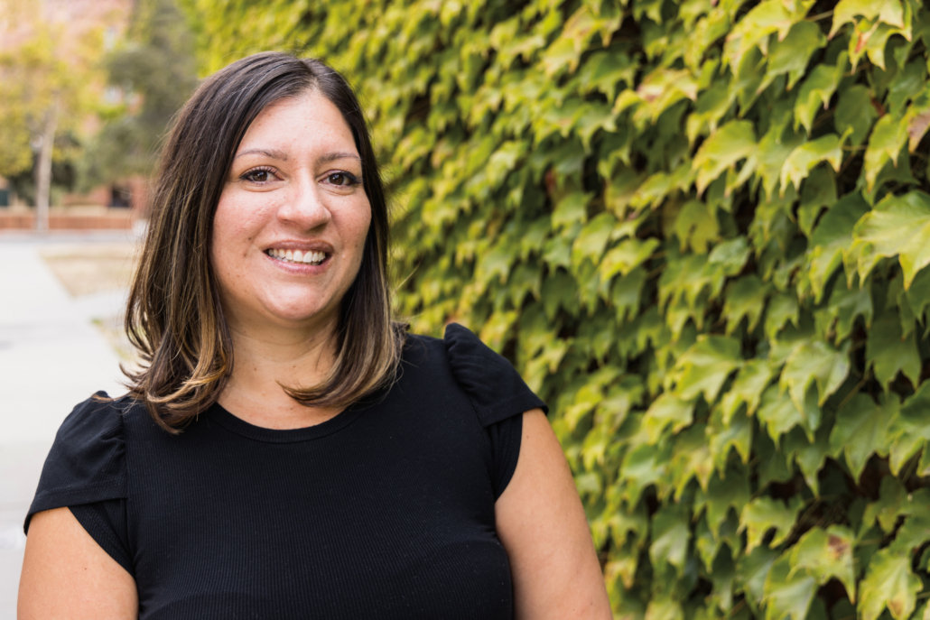 Melissa Villa Nicholas Headshot Professional headshot of a smiling person with shoulder length hair and a black shirt. They are standing in from of leafy green foliage.