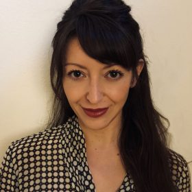 Michell_Kalani-280x280-1 Headshot of a person with long dark hair styled half up and side-swept bangs, wearing a black-and-white patterned blouse and dark lipstick, looking at the camera against a plain light-colored background.