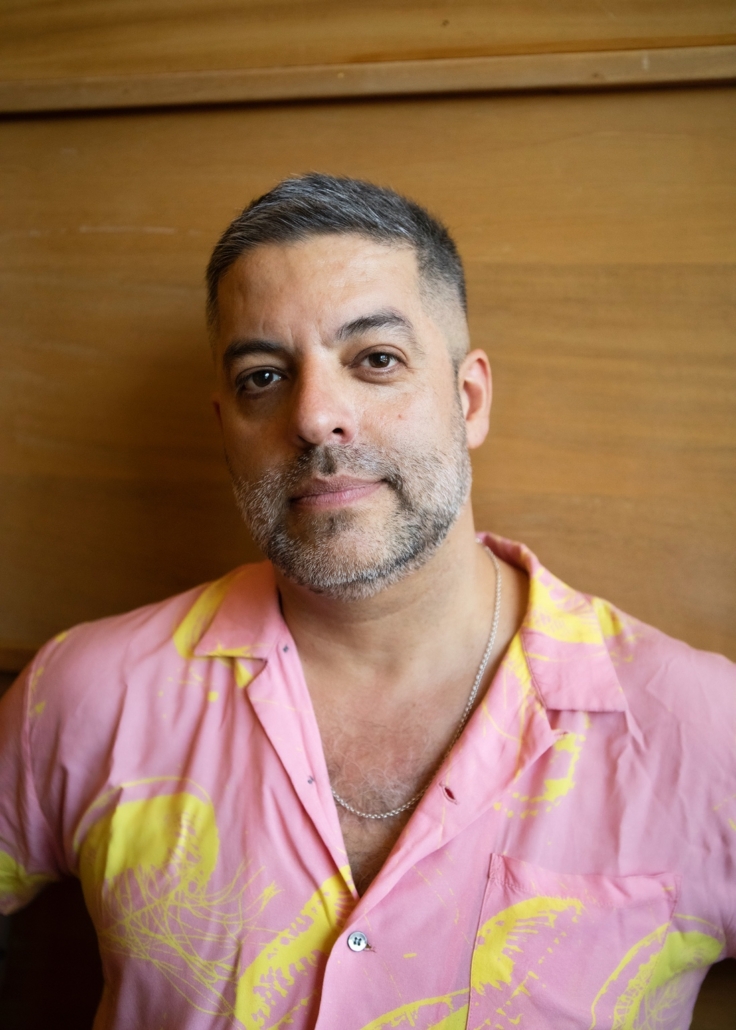 Miguel Gutierrez A man with short salt-and-pepper hair and a graying beard wears an open pink shirt with a yellow print and a silver chain necklace, photographed against a wood-paneled wall.
