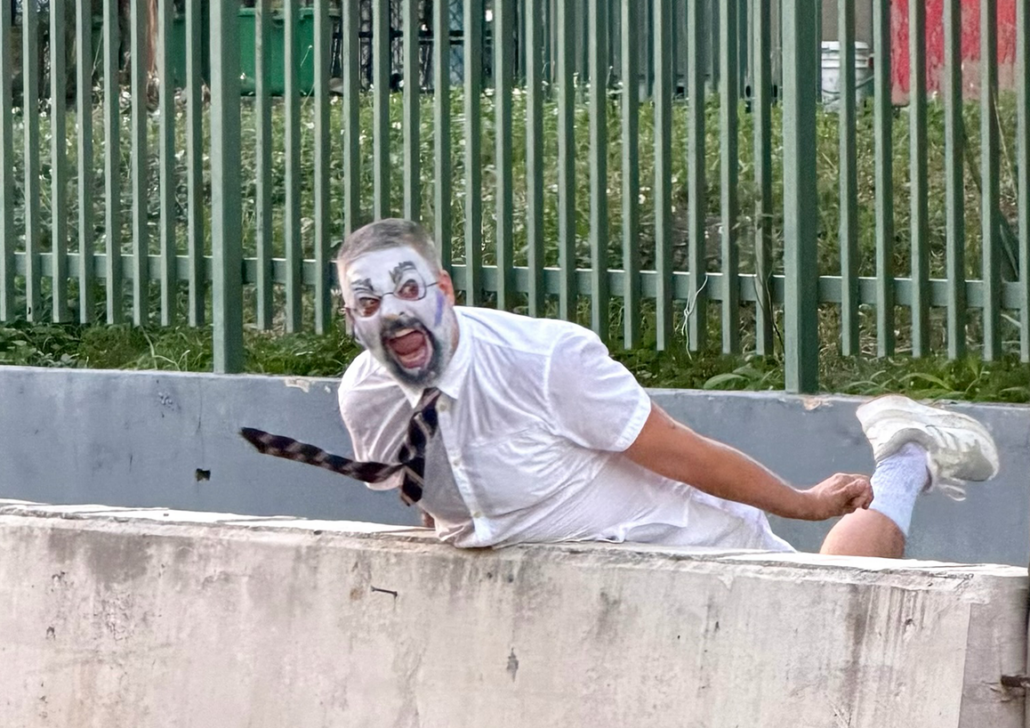 Miguel Gutierrez PROJECT A man with white and black face paint and glasses, dressed in a white shirt and tie, leans dramatically over a concrete barrier with mouth open in an exaggerated shout, one arm and leg stretched behind him, in front of a green metal fence.