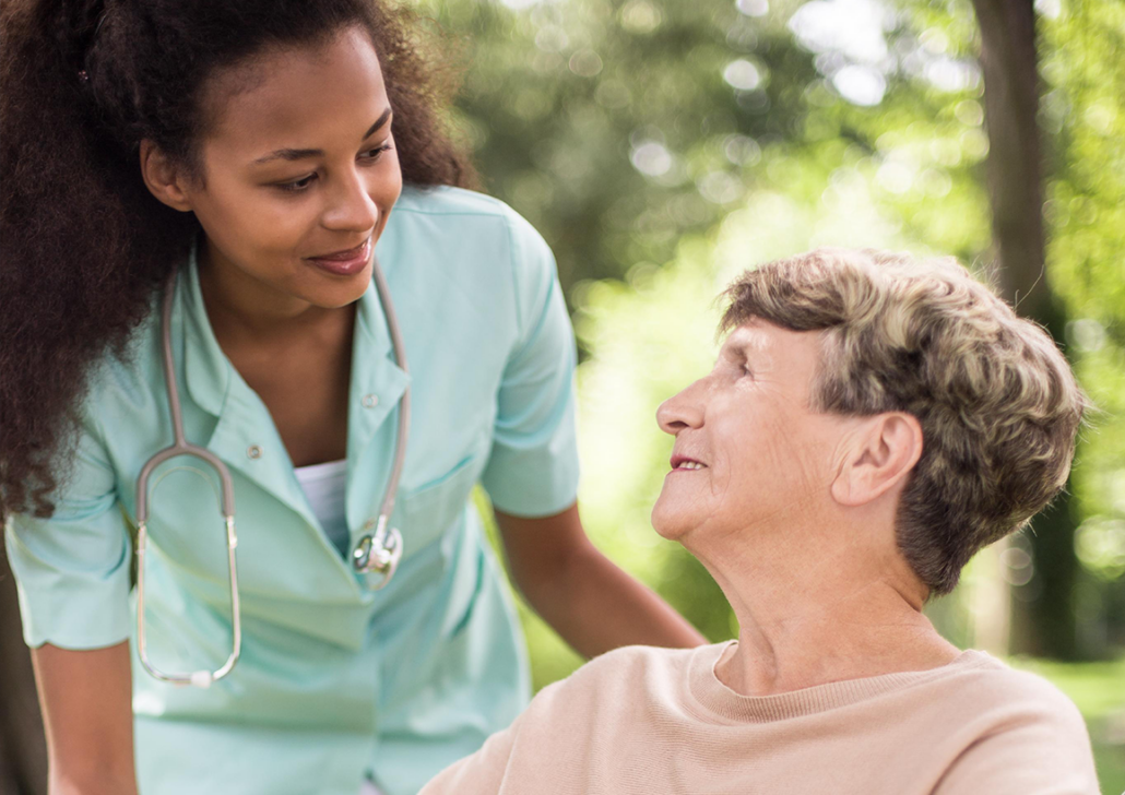 Paid Caregiving in Institutional Long-Term Care A caregiver in teal scrubs with a stethoscope leans down and smiles warmly at an older woman seated outdoors, who looks up and smiles back, surrounded by greenery.