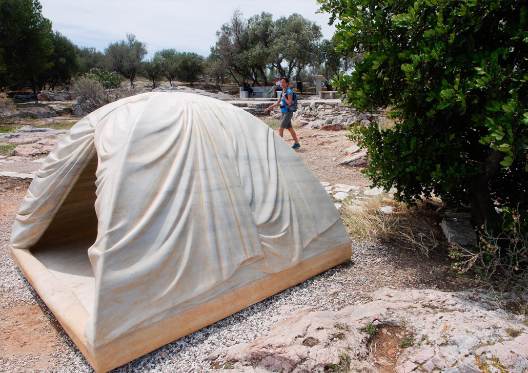 Shelter as Form: Rebecca Belmore's Marble Tent Grey tent in the wilderness. A person walks behind the tent and to the right of it is greenery.