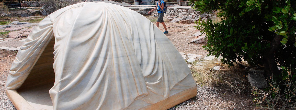 Grey tent in the wilderness. A person walks behind the tent and to the right of it is greenery.