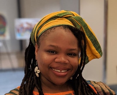 Shamara Wyllie Alhassan Picture A smiling woman with locs wears a yellow and green head wrap and floral earrings, photographed indoors against a softly blurred background.