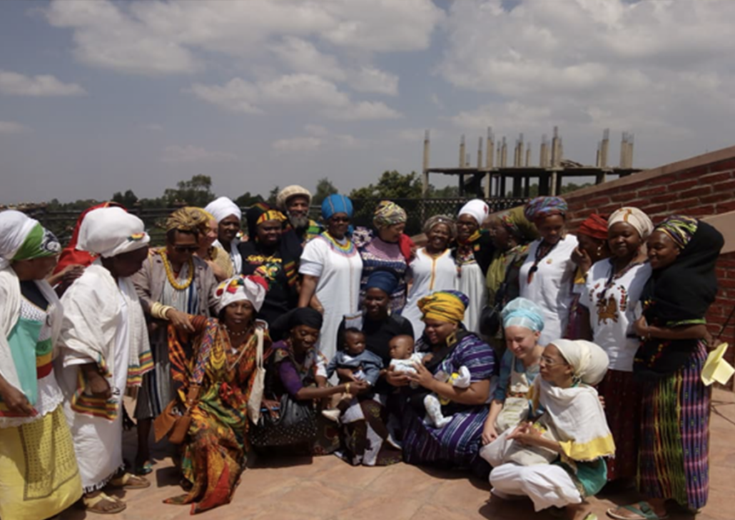 Shamara Wyllie Alhassan Project Photo A large group of men, women, and children in colorful traditional dress and head wraps pose together on a rooftop terrace, with two babies held in the front row and a building under construction visible in the background.