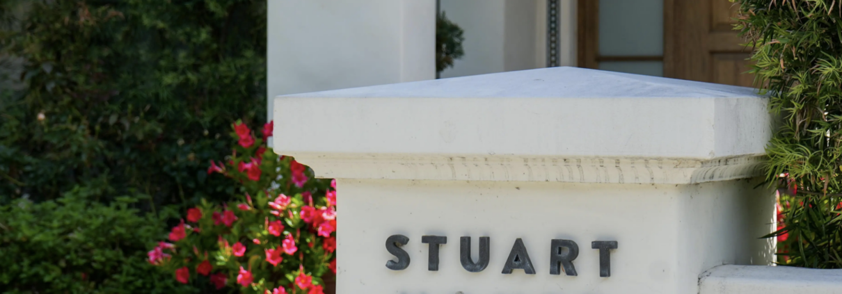 White stucco entrance pillar with the words “Stuart House” in dark metal letters, in front of a wooden door, with red-pink flowering bushes and green shrubs alongside.