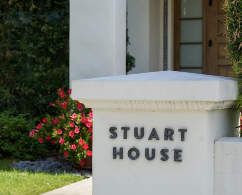 White stucco entrance pillar with the words “Stuart House” in dark metal letters, in front of a wooden door, with red-pink flowering bushes and green shrubs alongside.