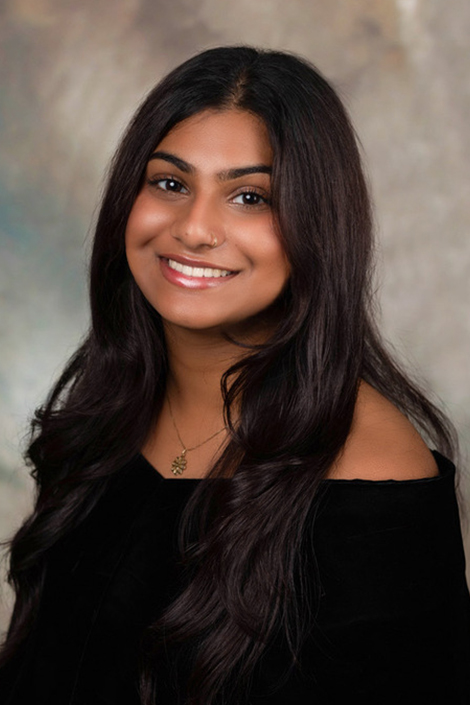 Portrait of a young woman with long, wavy dark brown hair, smiling warmly at the camera. She wears a black off-the-shoulder top, a delicate gold pendant necklace, and a small nose stud. The background is a soft, painted-style backdrop in muted grays, tans, and hints of blue.